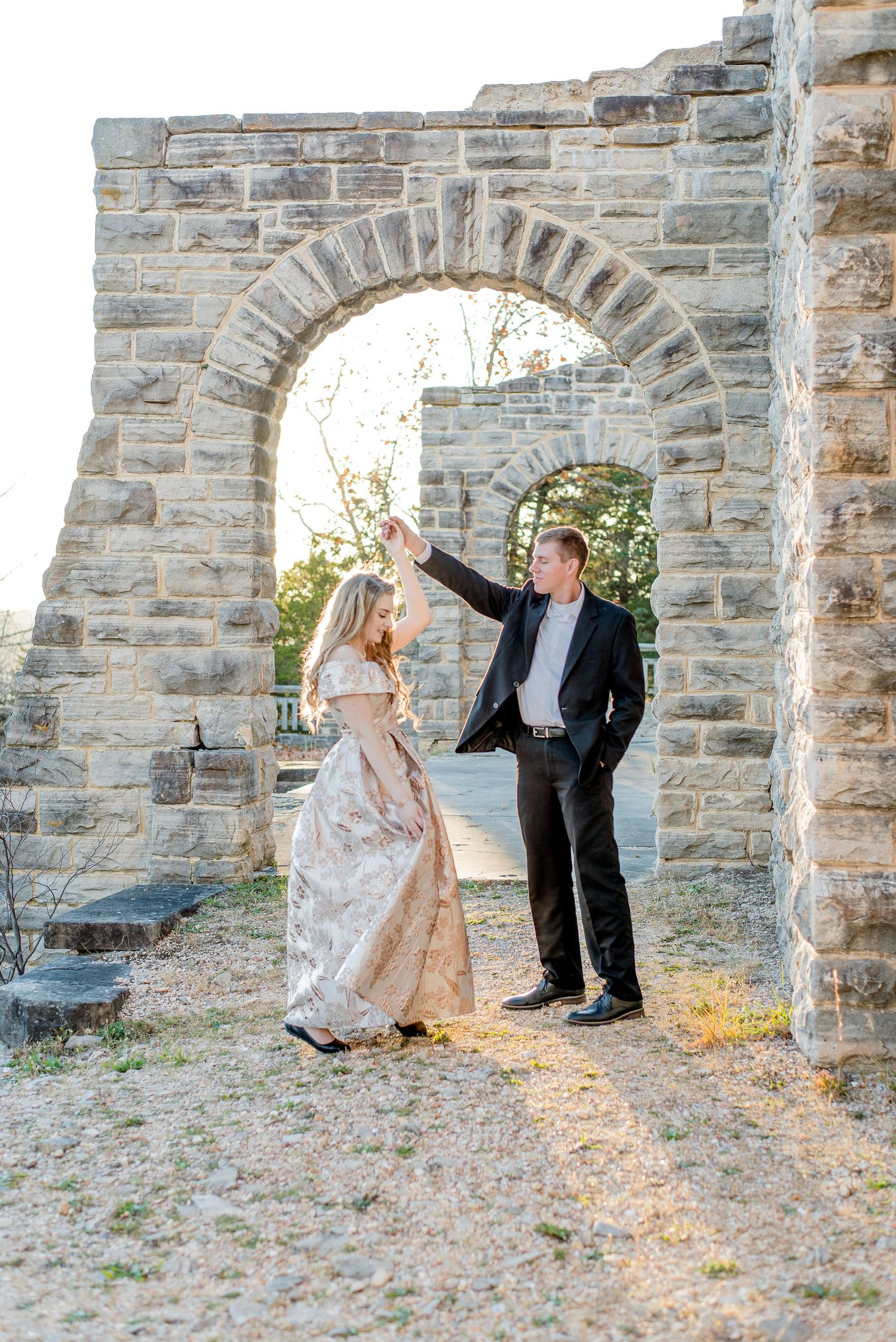 A couple in formal attire dance together under a stone archway during sunset at a historic ruins location.