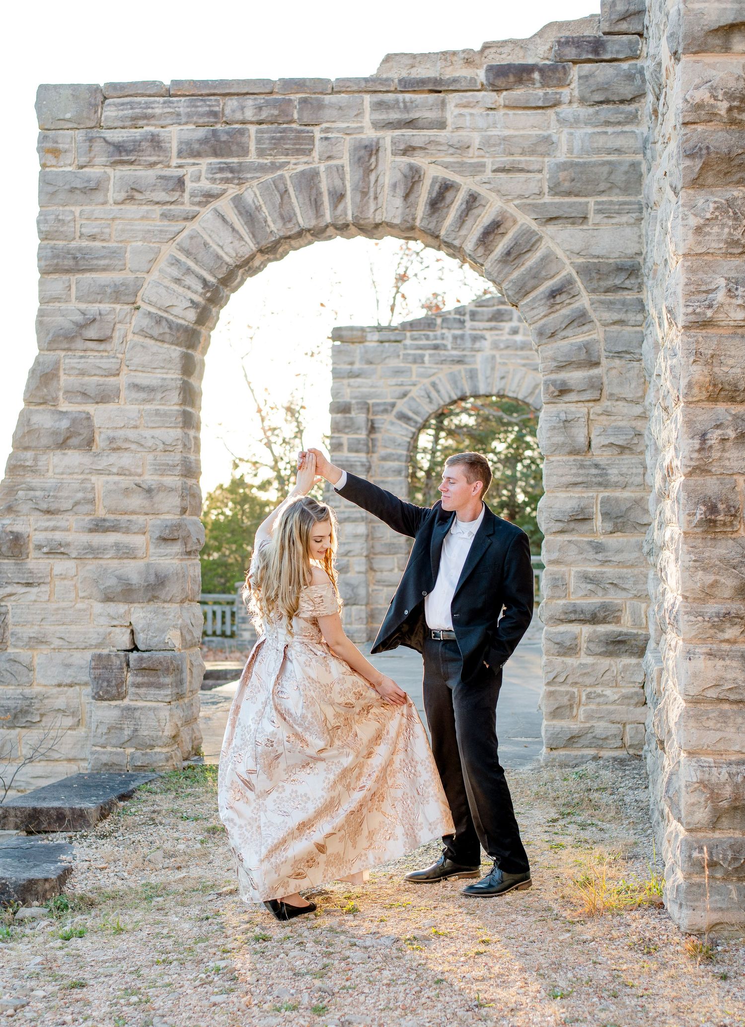 A romantic scene with a couple dancing under a stone archway at sunset wearing formal evening attire.