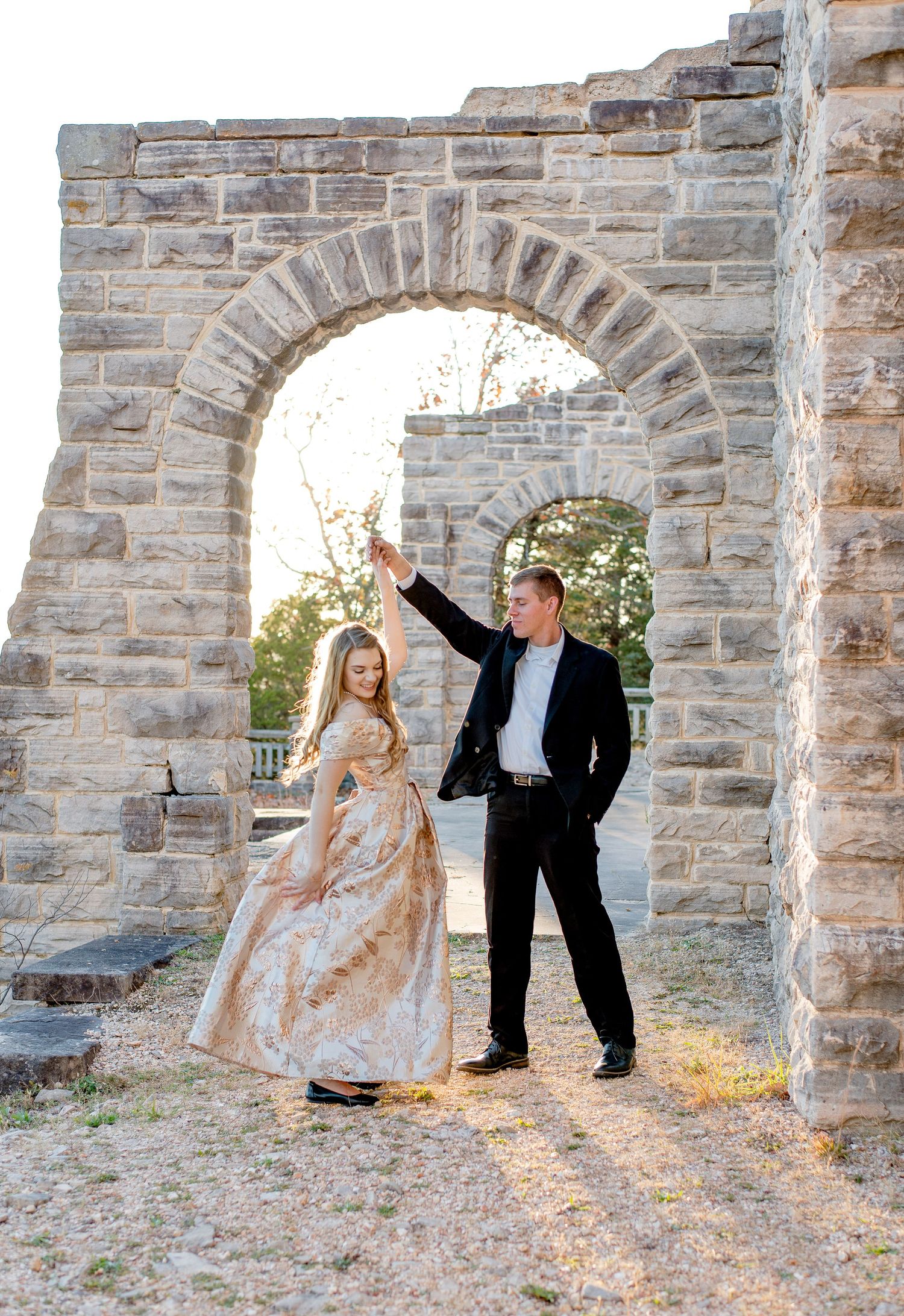 Couple in formal attire poses under stone archway at sunset in romantic medieval-style setting.