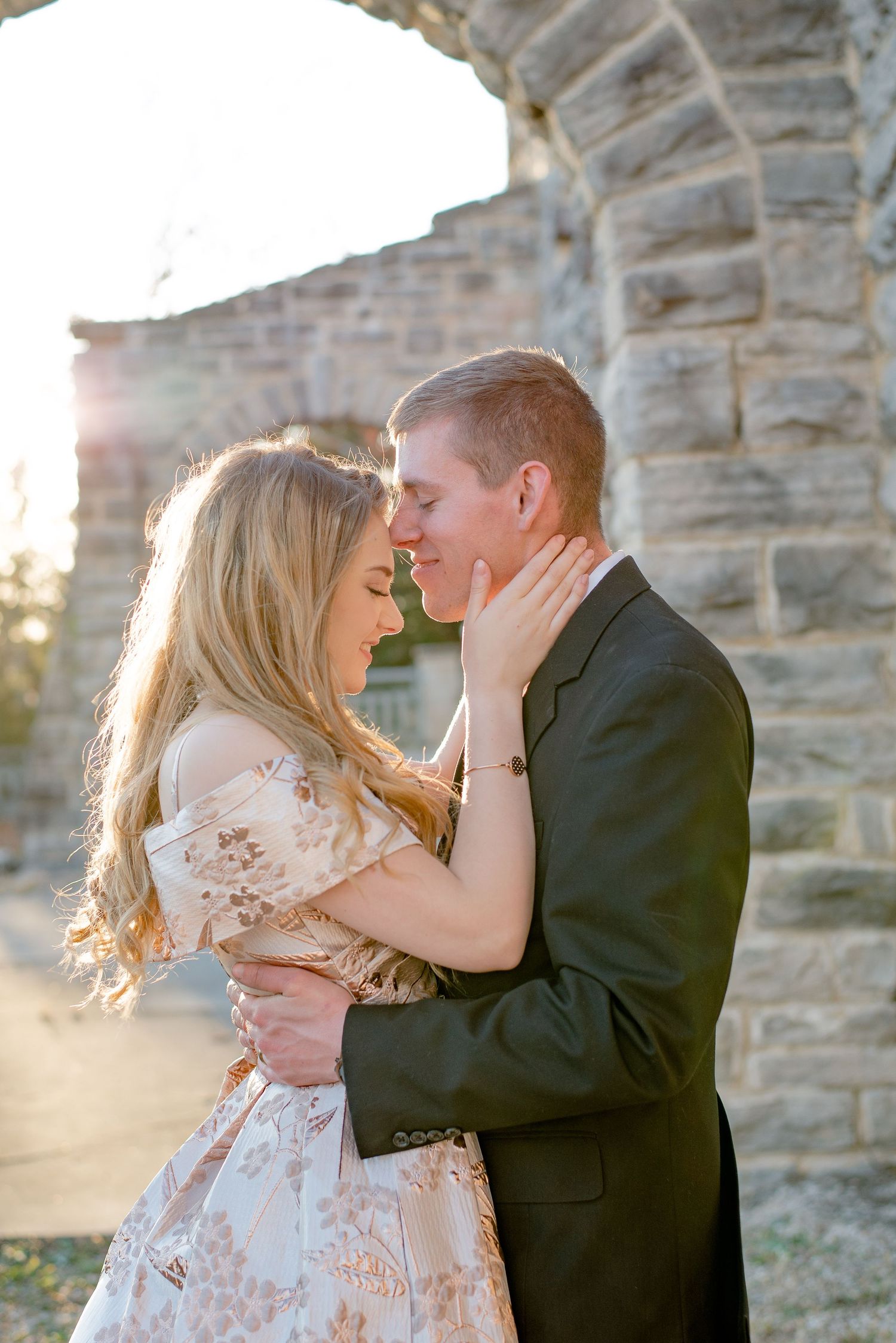 A romantic couple embraces in front of a stone wall during golden hour sunset with the sunlight creating a warm glow.