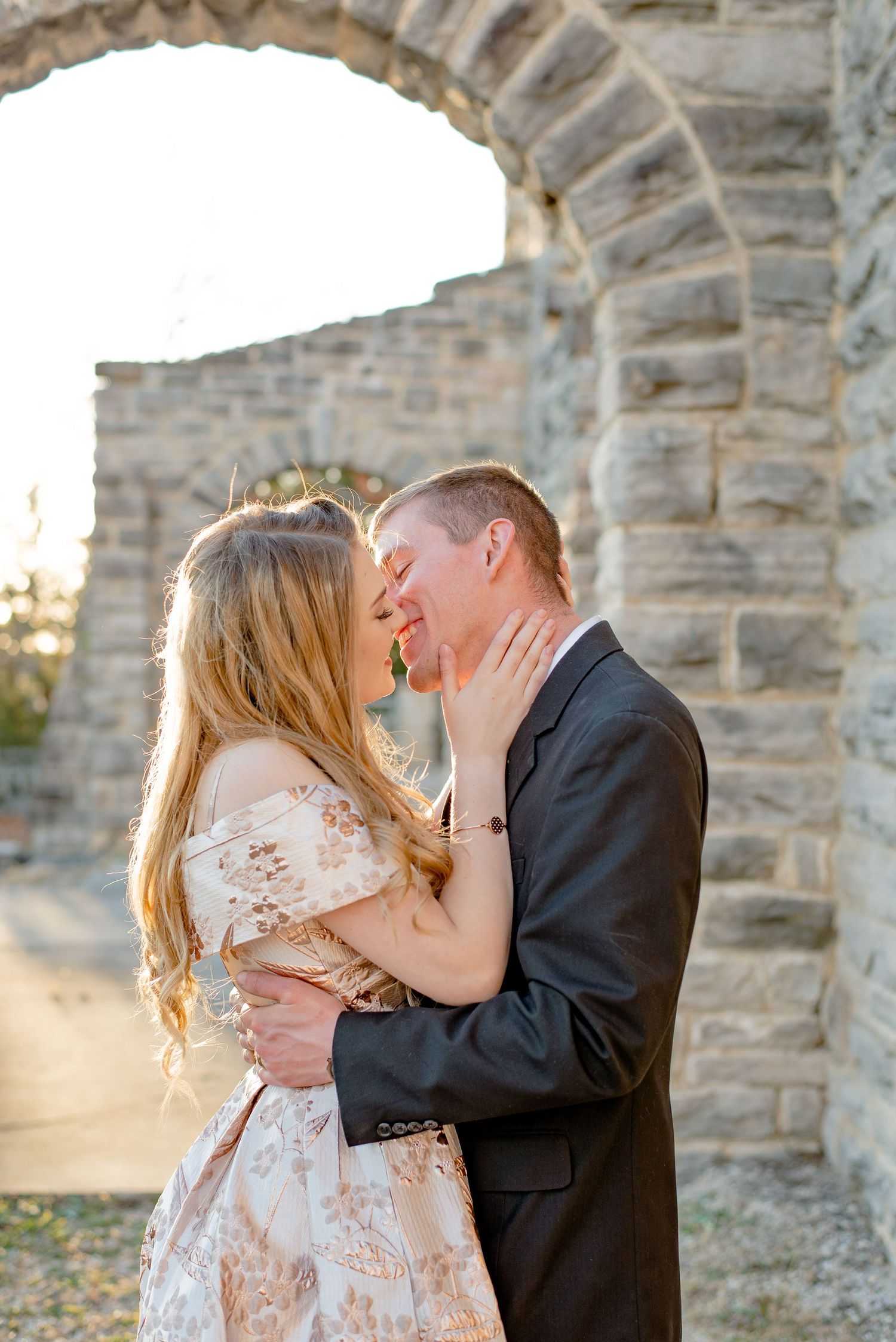 A romantic couple embraces under stone archways at sunset wearing formal attire and a blush lace dress.