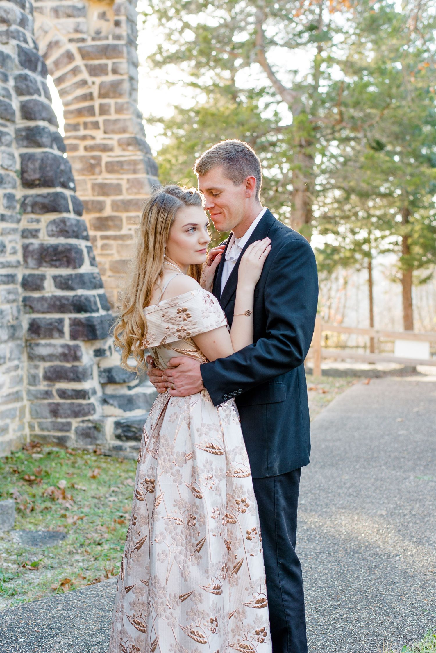 A romantic scene of a couple embraces outdoors near a stone building wearing formal attire.