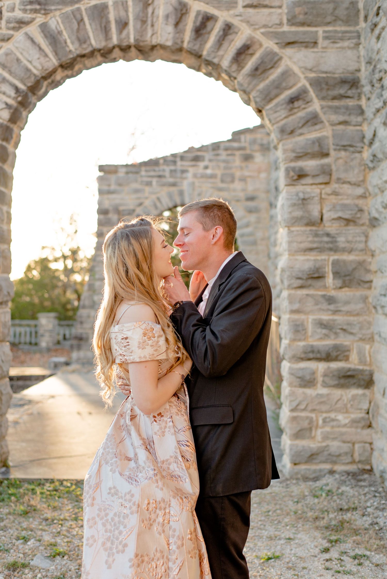 Romantic couple embraces under stone archway at sunset during formal photo session.