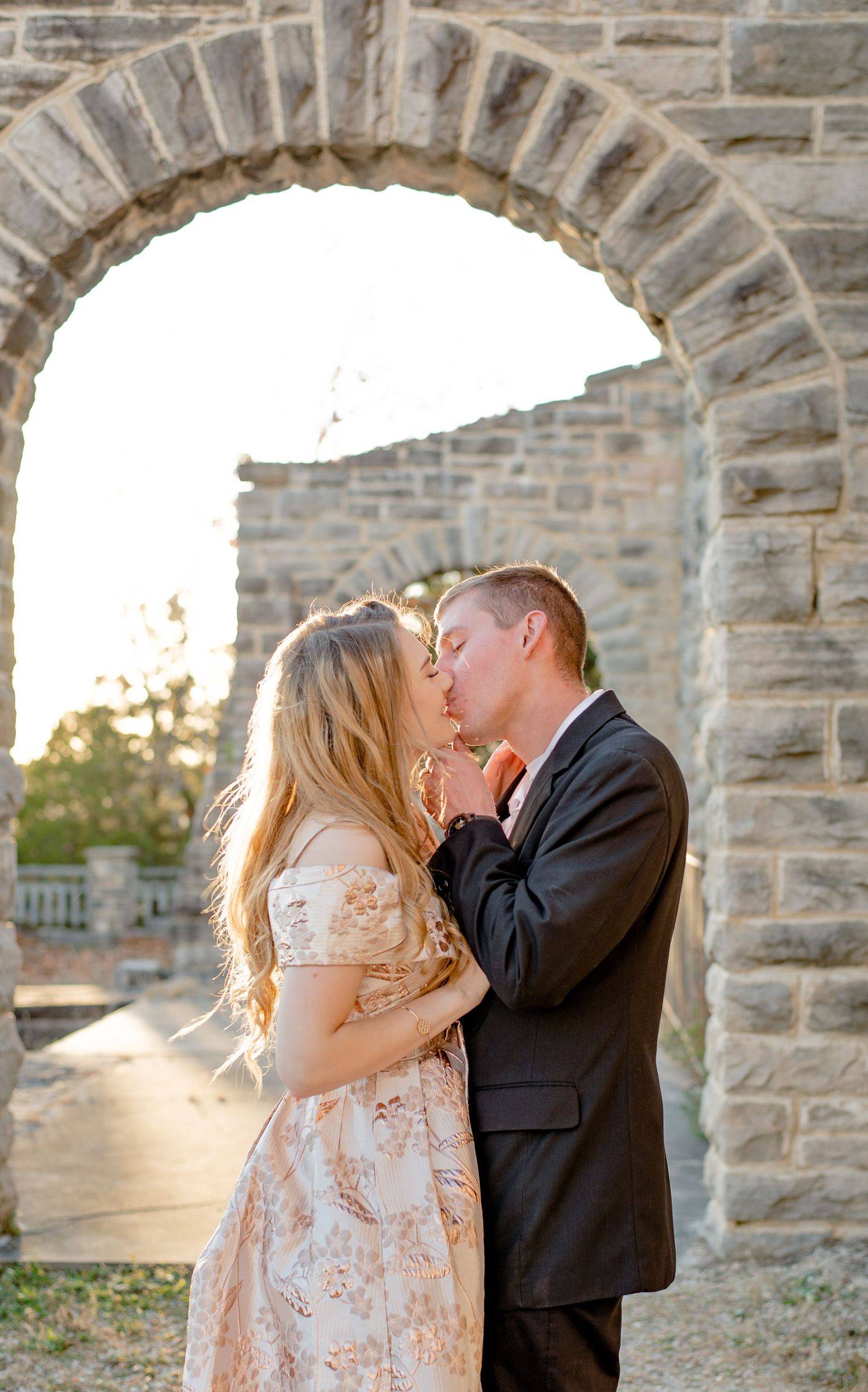 Romantic couple sharing an intimate kiss under a stone archway at sunset with golden light.