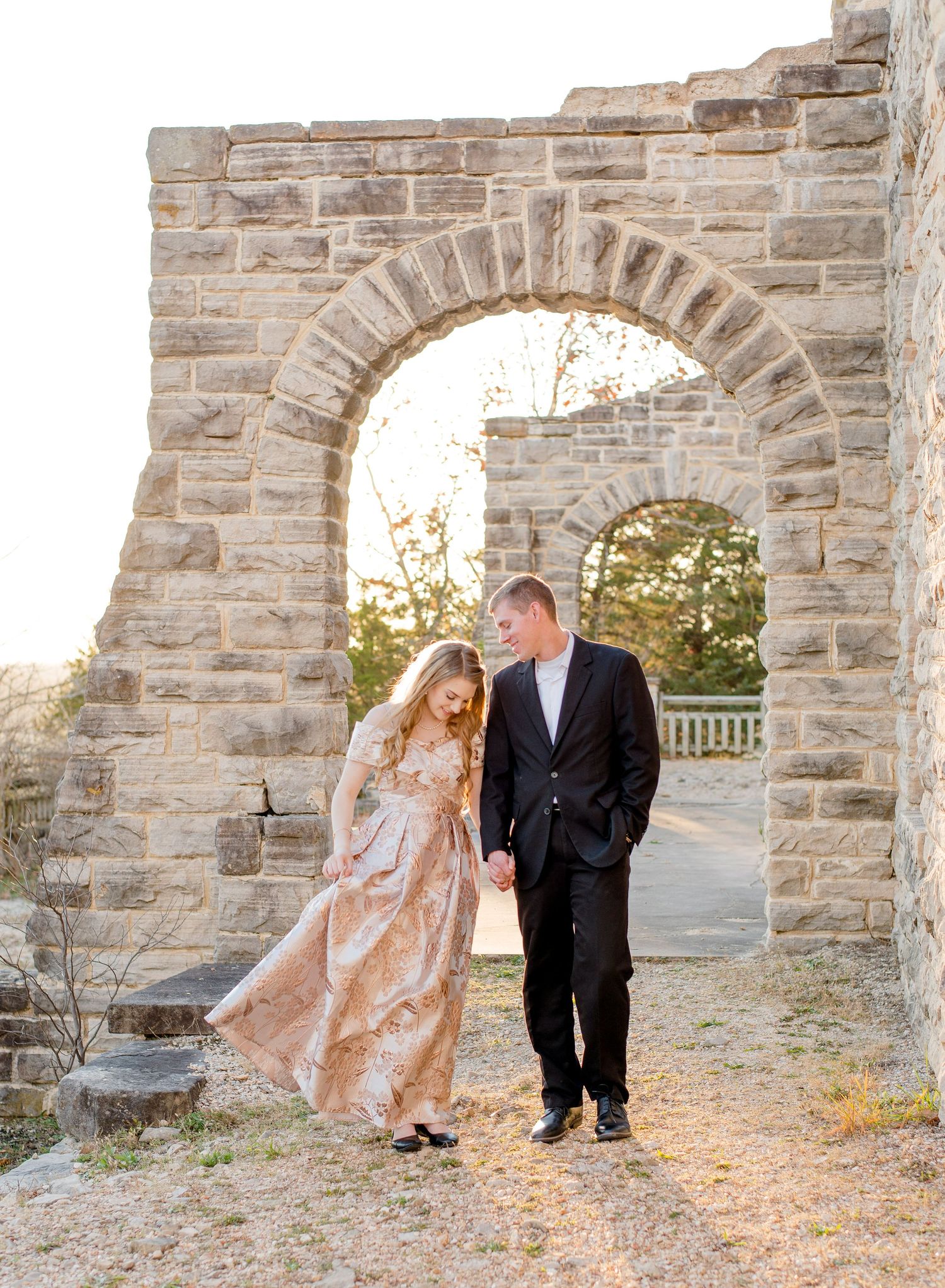 A couple stands together under a rustic stone archway at sunset in elegant formal attire.