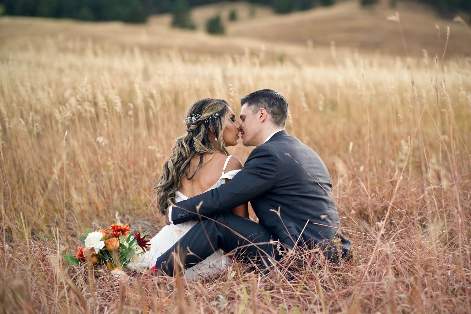 Romantic couple sitting in tall golden grass with wedding bouquet at sunset.