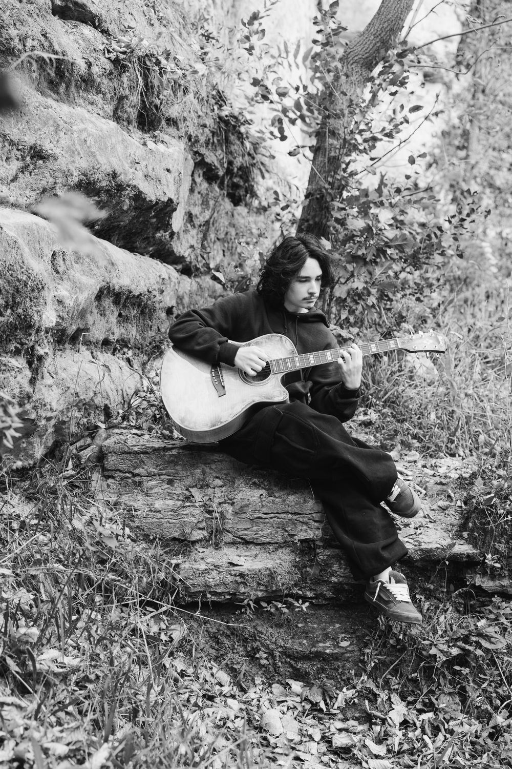 Black and white photo of musician playing acoustic guitar on rocky outcrop.