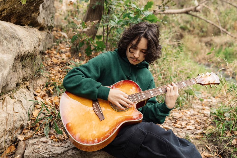Musician in green sweater plays acoustic guitar while sitting on fallen log in nature setting.