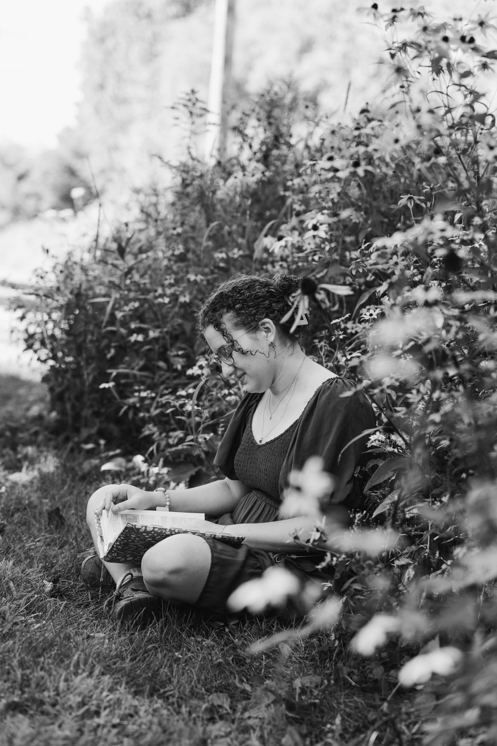 Black and white photo of someone reading outdoors among flowering bushes.