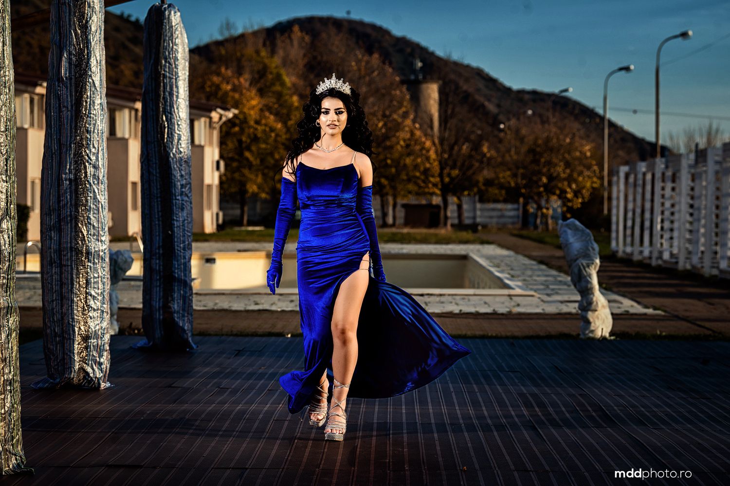 Model in flowing royal blue evening gown with high slit walking on outdoor pathway at sunset.