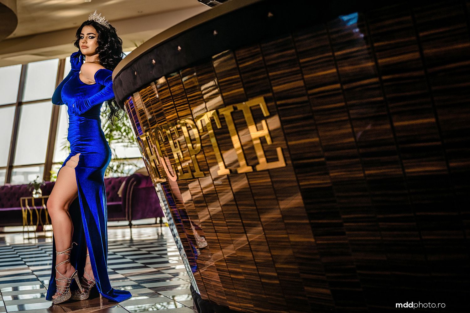 A figure in a vibrant royal blue evening gown with a side slit poses next to a curved wood-paneled wall.