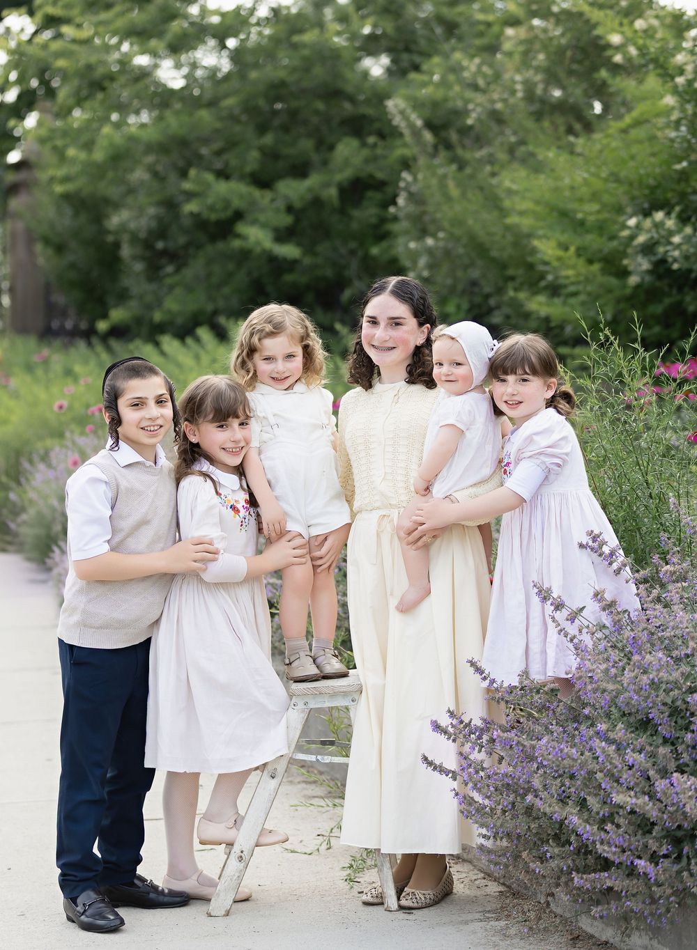 A group in coordinating white and cream outfits poses together near blooming purple wildflowers on a garden path.