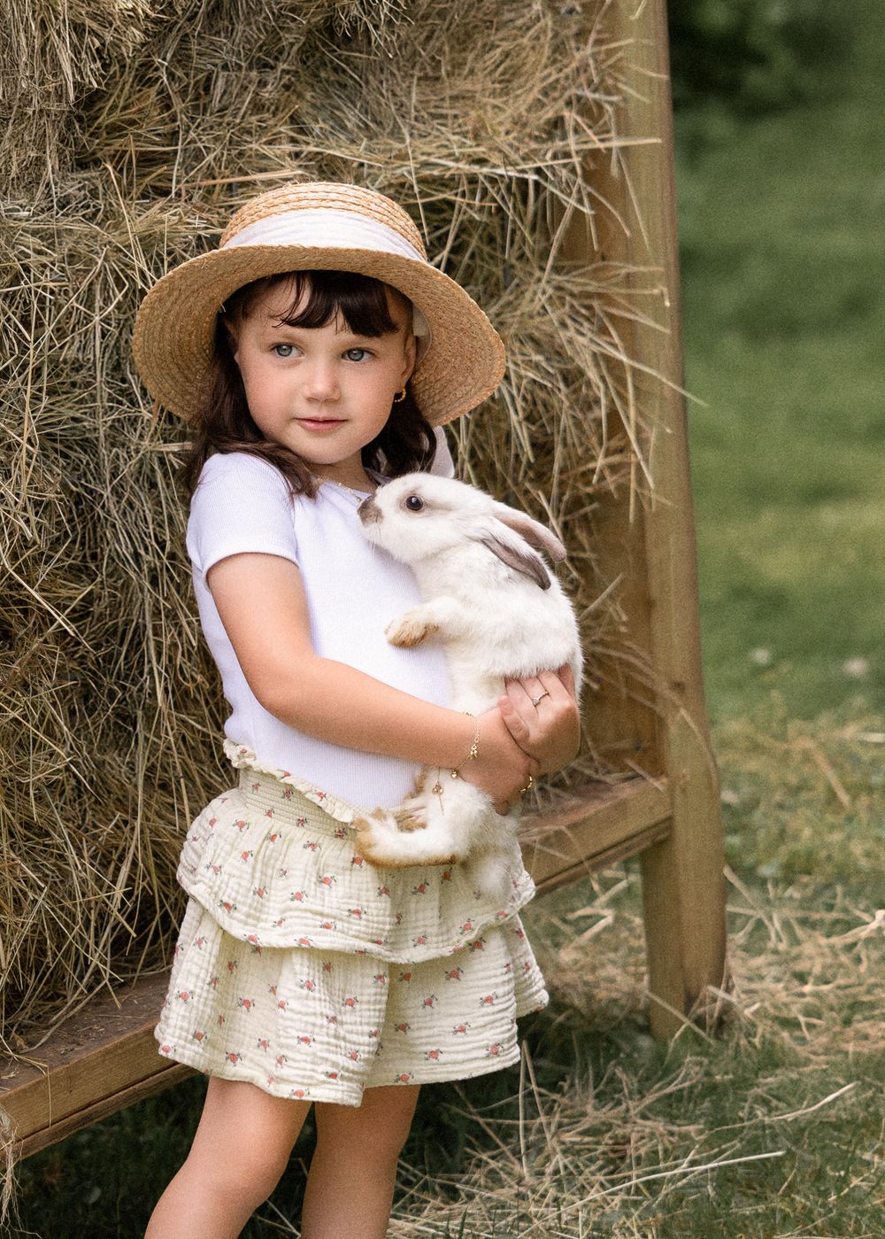 A young child in a straw hat and white dress holds a fluffy white bunny rabbit in front of a rustic wooden fence.