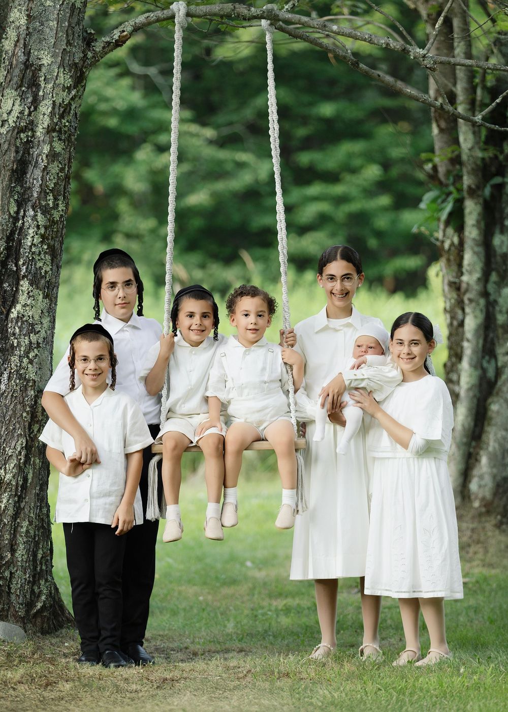 Group of children in white clothing posing outdoors near a tree swing in a lush green setting.