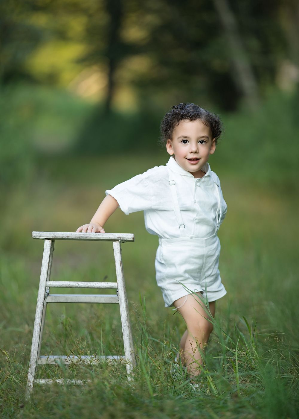 Young boy in white outfit standing beside a rustic white stool in a lush green outdoor setting.