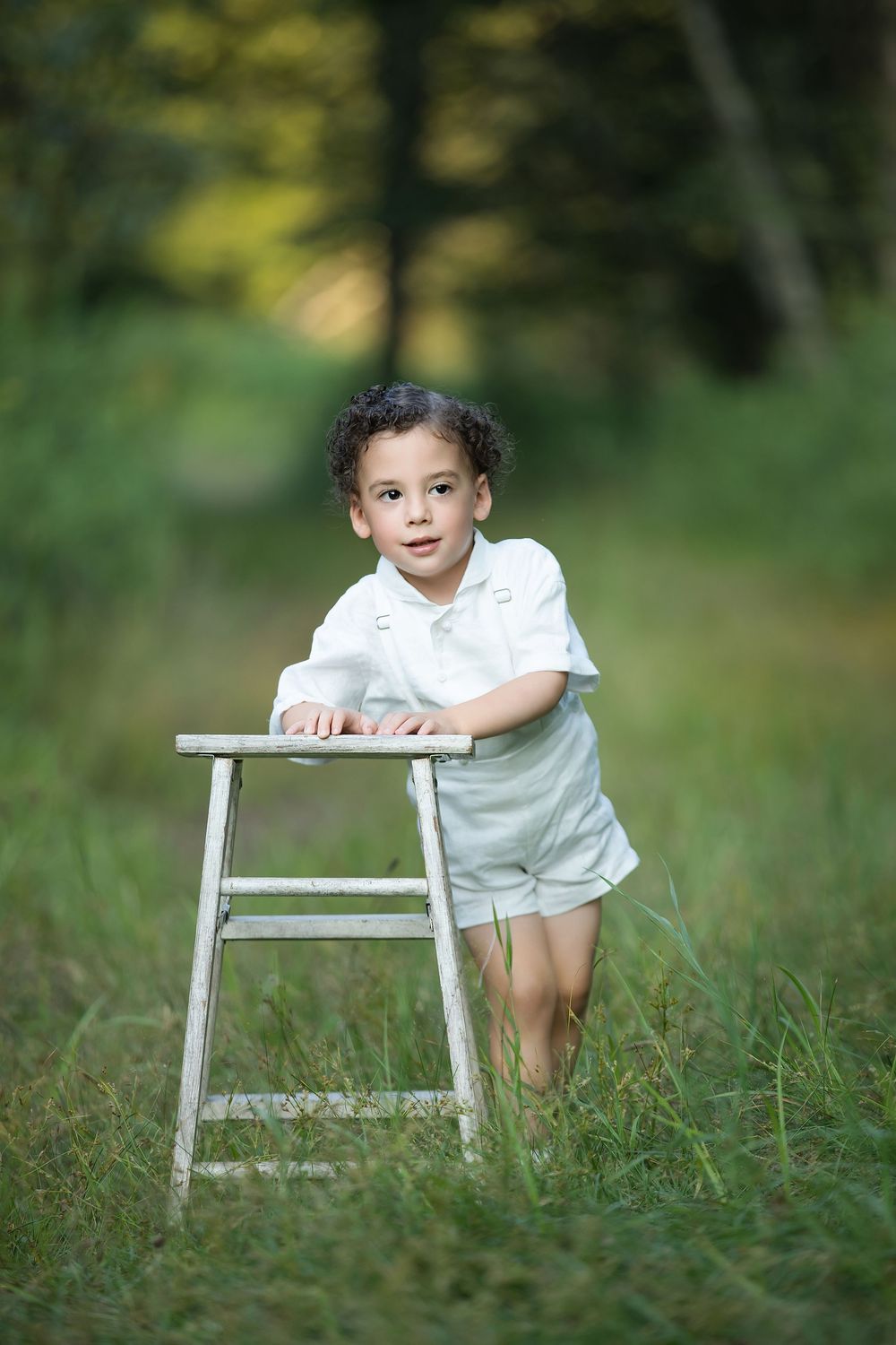 Young boy in white outfit leaning on a rustic wooden stool in a lush green outdoor setting.