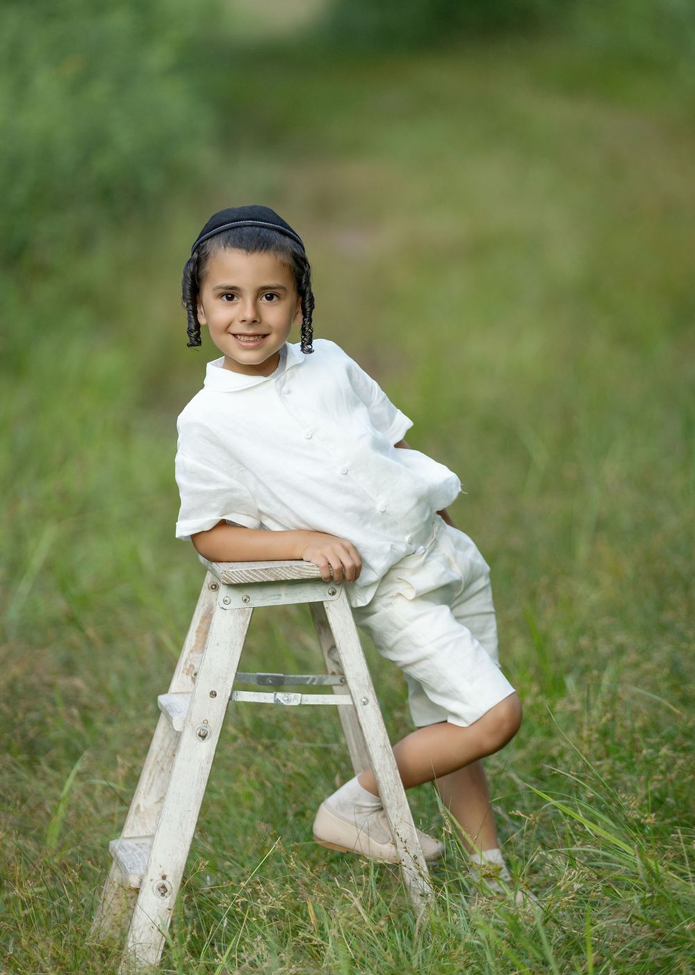 A smiling child in white outfit sits on a wooden stepladder in a lush green field.