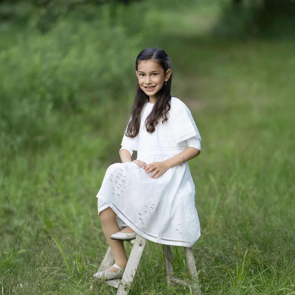 A young girl in a white dress sits on a wooden stool, smiling outdoors in a lush green garden.