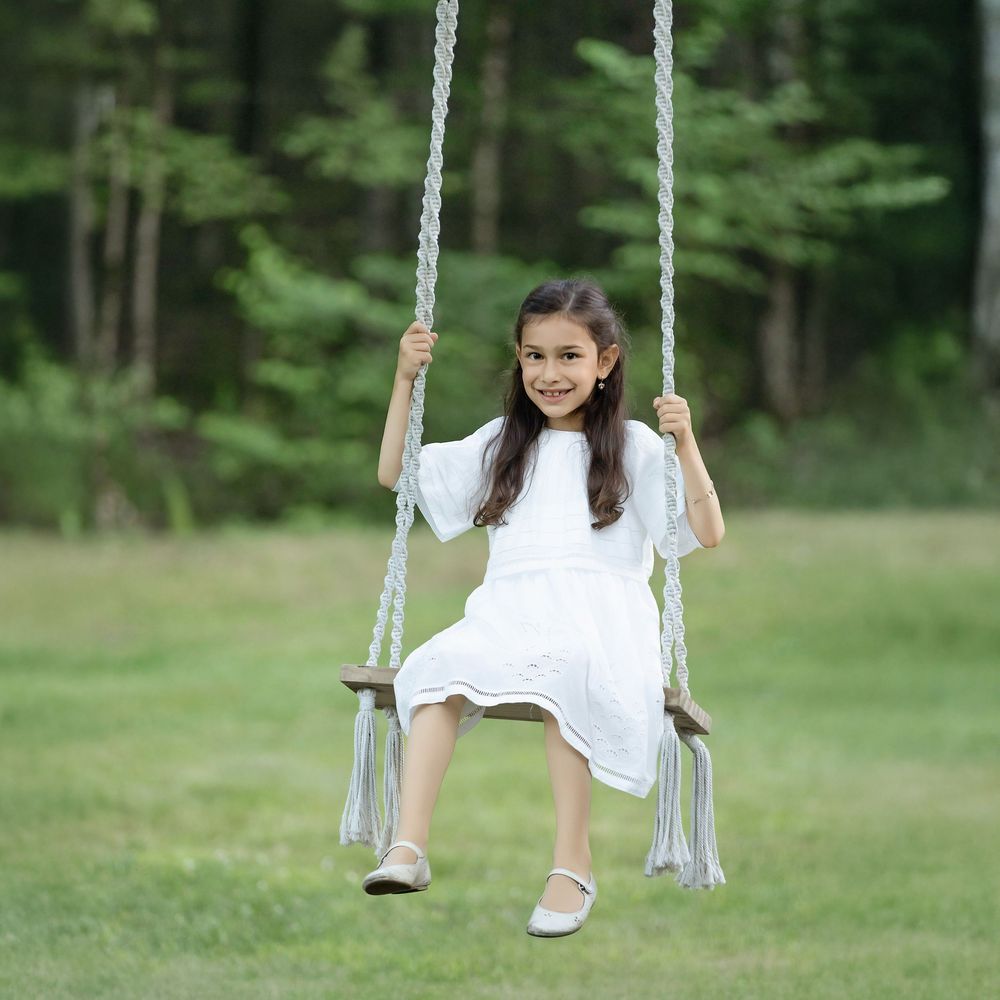 Young girl in white dress smiling on a wooden rope swing in a lush green park setting.