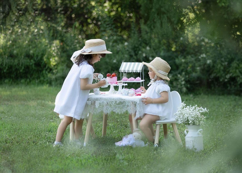 Two children in white dresses and sun hats enjoy an elegant outdoor tea party setup in a lush garden setting.