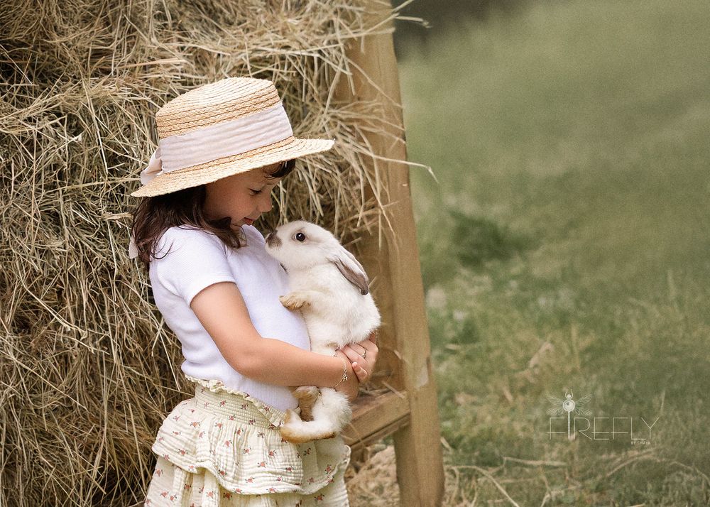 A person in a sun hat holds a white bunny rabbit against a rustic wooden and straw backdrop.