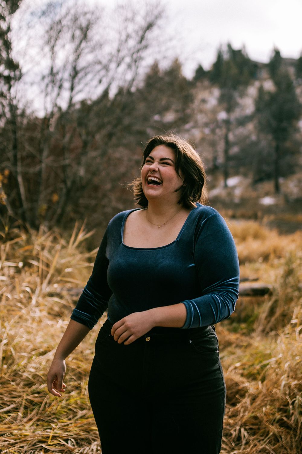 A person in a navy blue dress laughs joyfully while standing in tall autumn grass with mountains in the background.