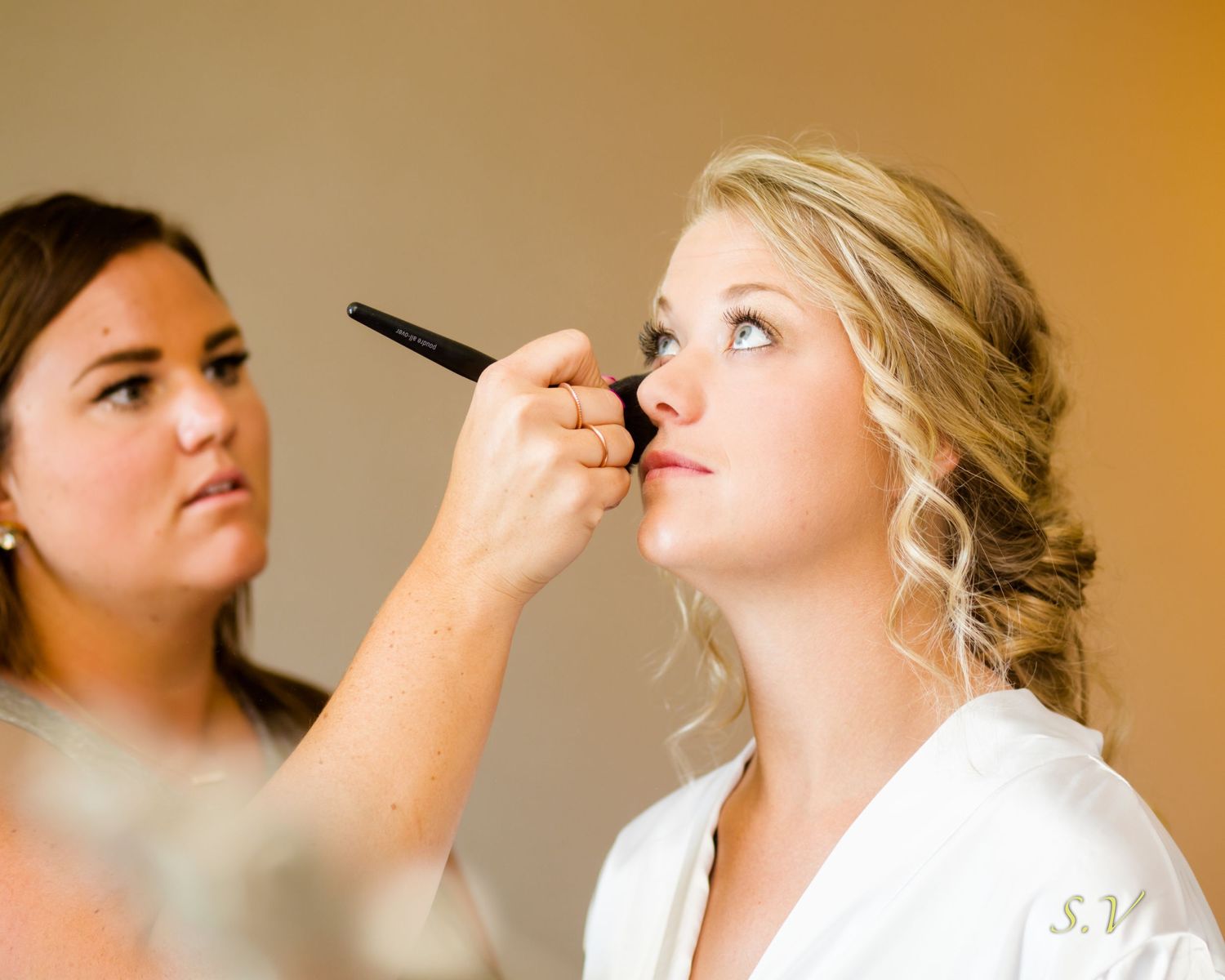 A makeup artist applies cosmetics to someone wearing a white robe during wedding preparations.