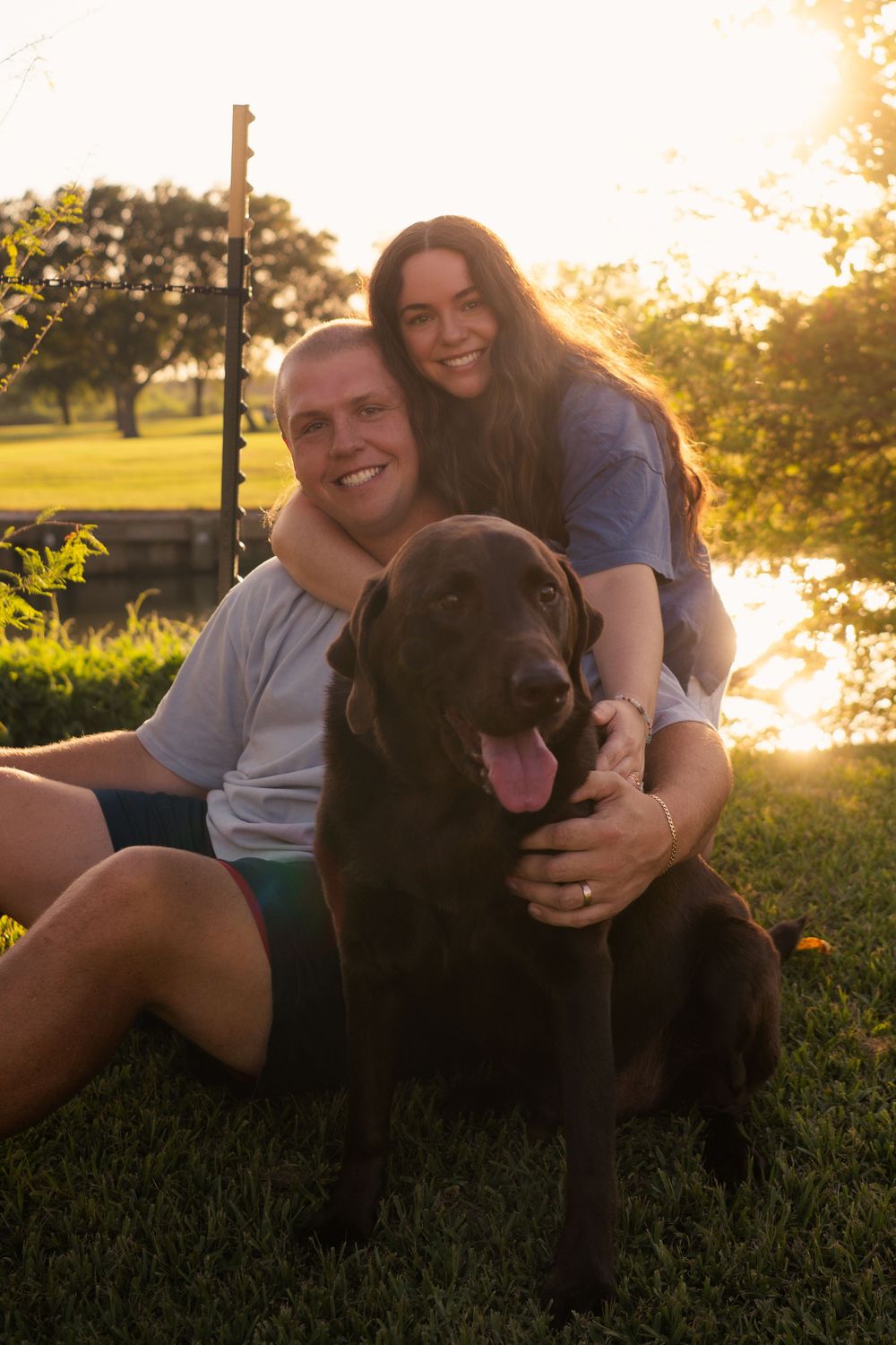 Smiling couple sitting on grass at sunset with a chocolate Labrador dog, backlit by golden hour light near a pond.