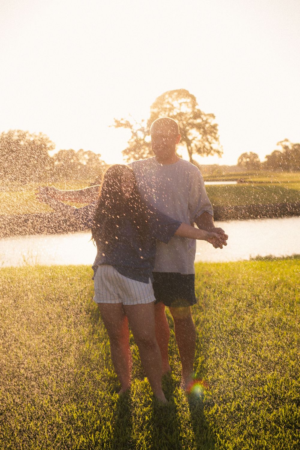 Couple embracing in golden sunset light near a pond, with water droplets sparkling around them on green grass.