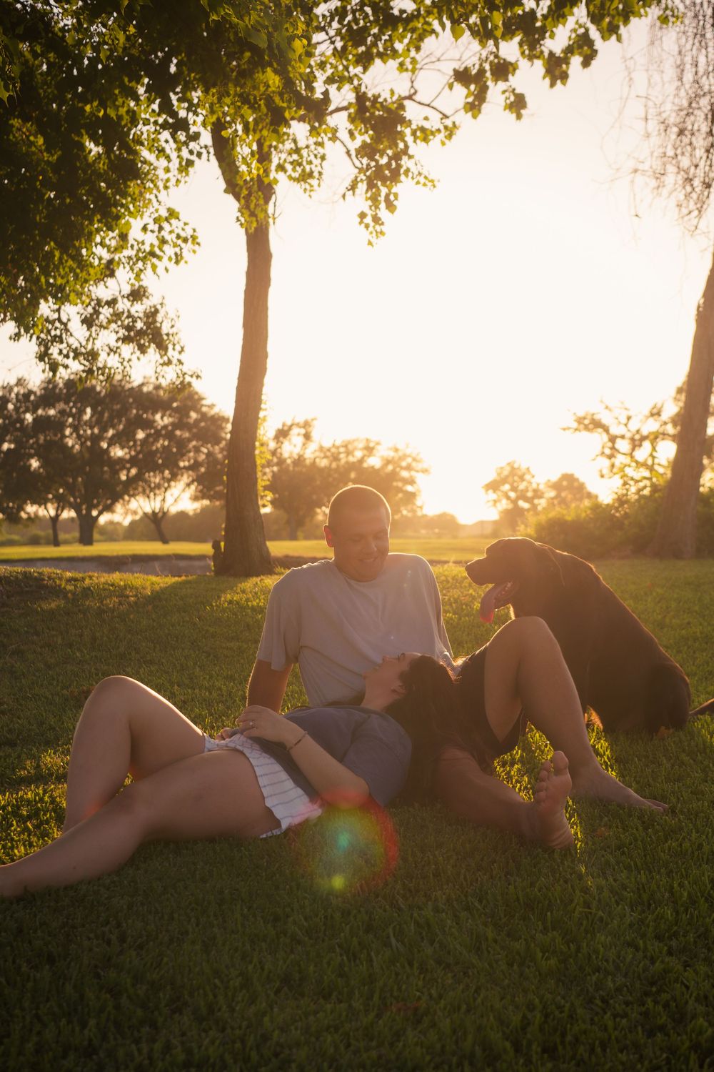 Couple relaxing on green grass with their black dog at golden hour sunset in a park.