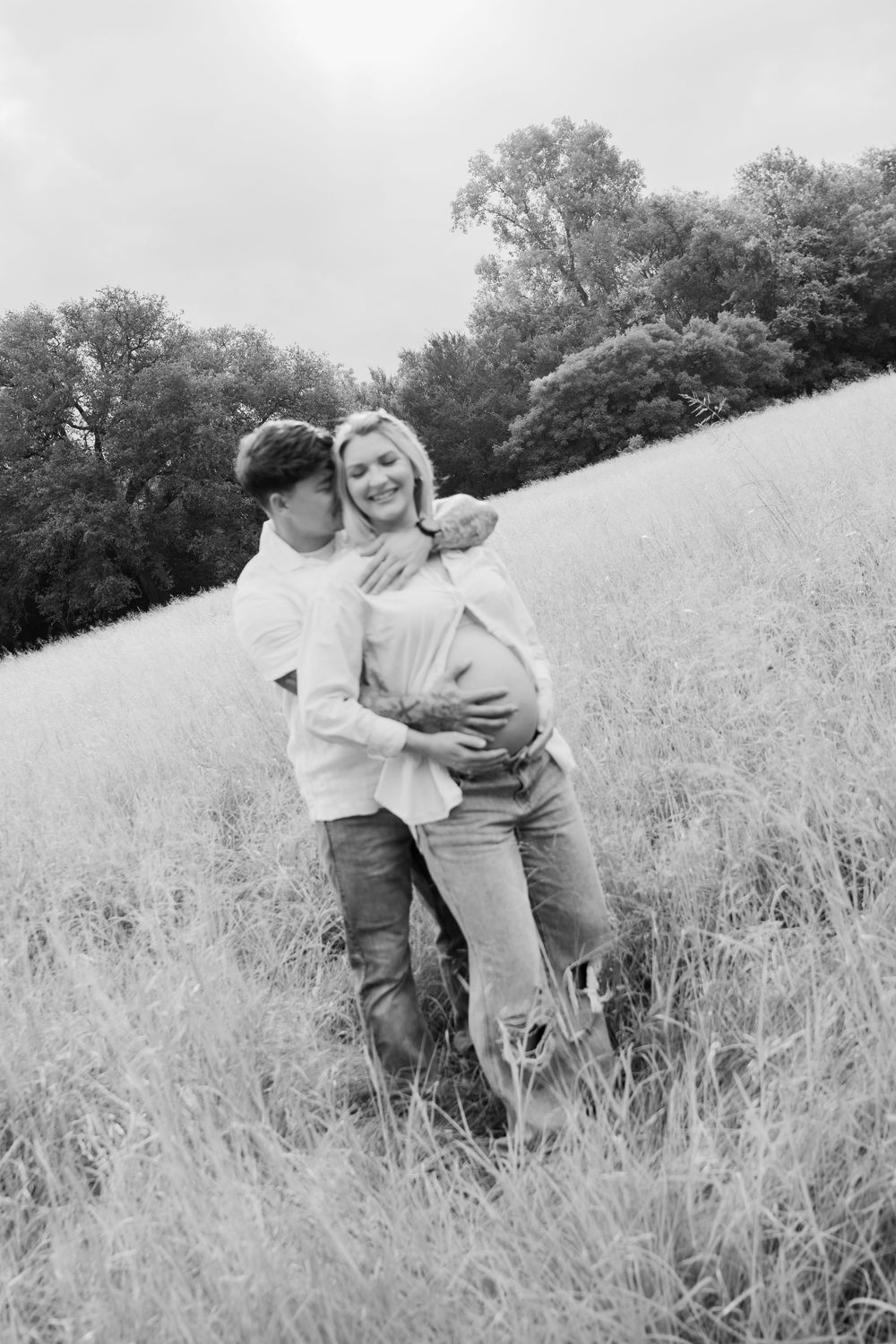 Black and white photo of a couple in a field, man embracing pregnant woman from behind, both smiling outdoors.