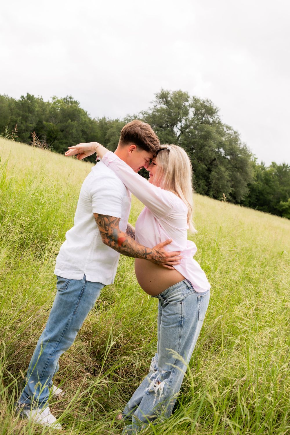 Pregnant couple embracing in a green field, man with tattoos in white shirt and jeans, woman in pink top showing baby bump.