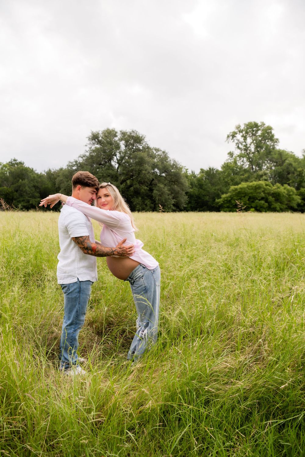 Couple embracing in a green field, pregnant woman in pink top and jeans, man in white shirt, trees in background.
