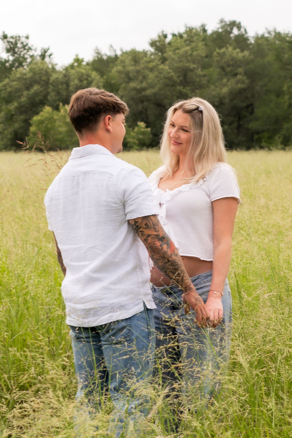 A couple in white tops and jeans hold hands in a lush green field, sharing a romantic moment outdoors.