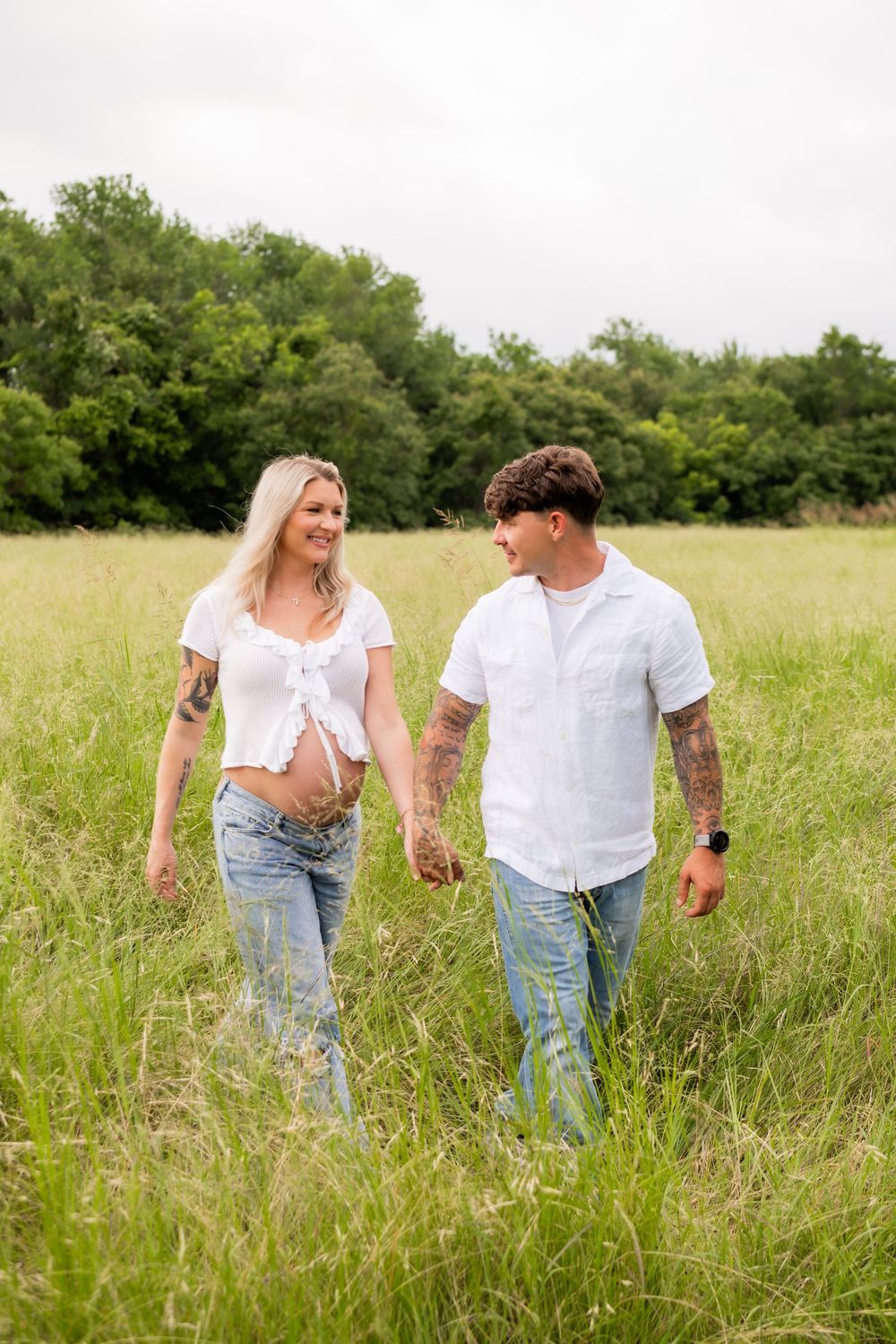 Pregnant couple holding hands walking through green field, woman showing baby bump in white crop top and jeans.