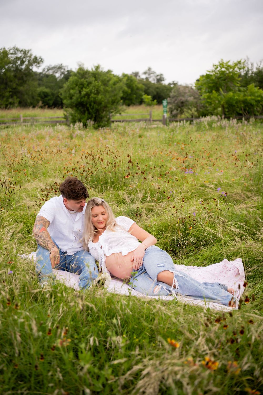 Pregnant couple relaxing in a green meadow, woman lying on man's lap showing baby bump, wearing casual jeans and white tops.