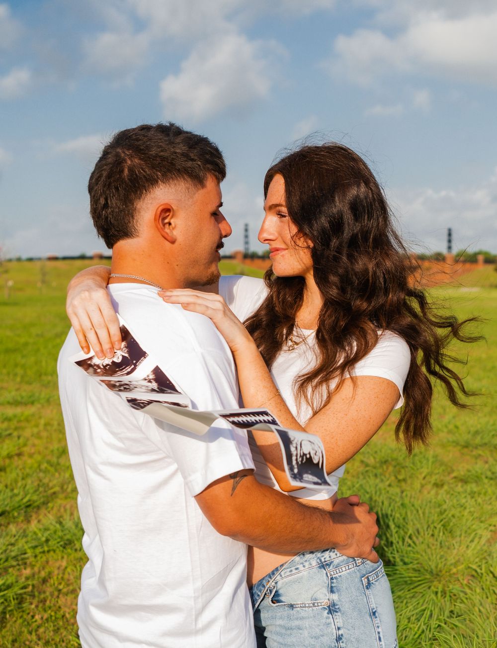 Couple in white shirts embracing outdoors, holding ultrasound photos, announcing pregnancy in a green field.