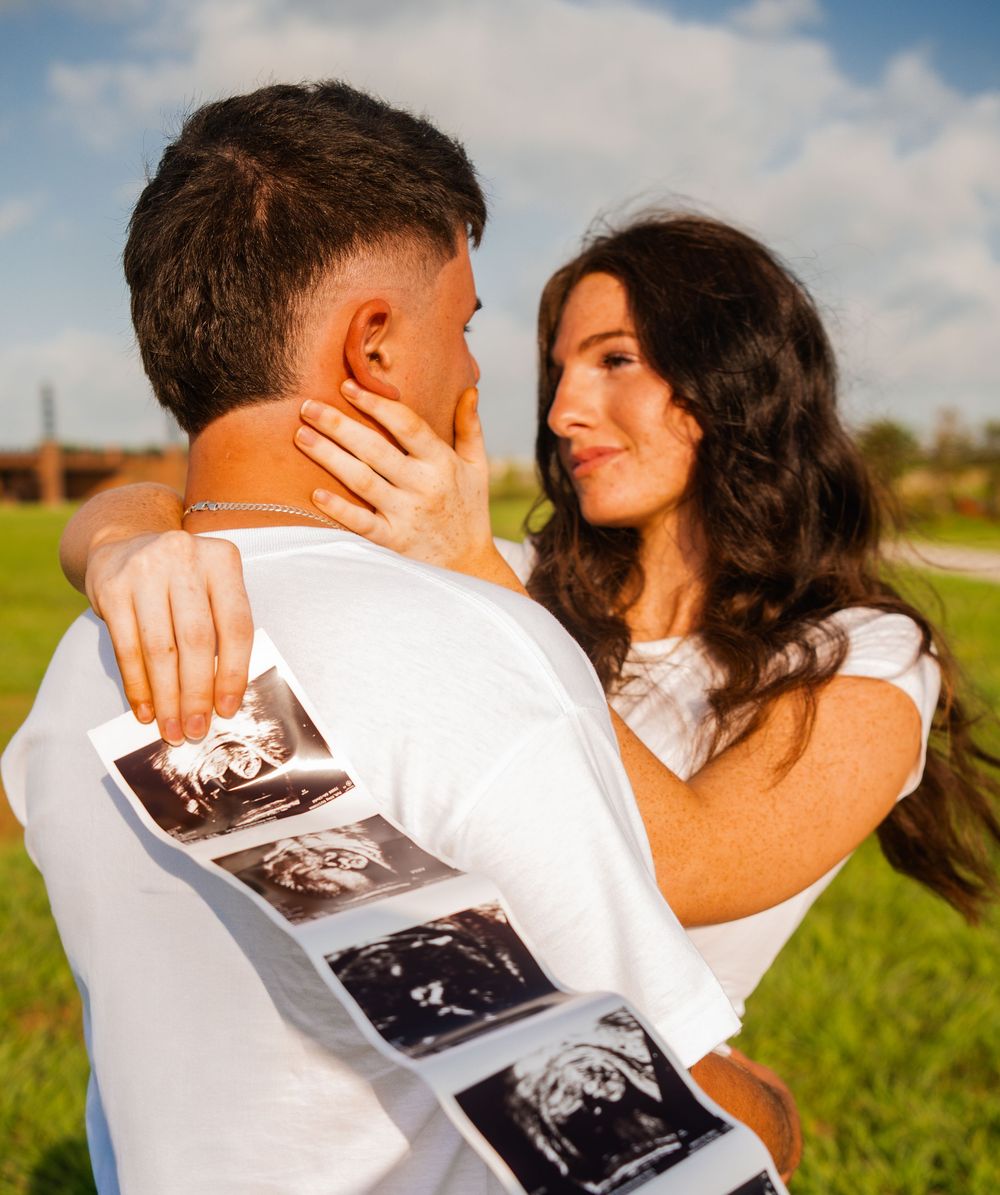 Couple embracing outdoors holding ultrasound photos, pregnancy announcement in sunny green field.