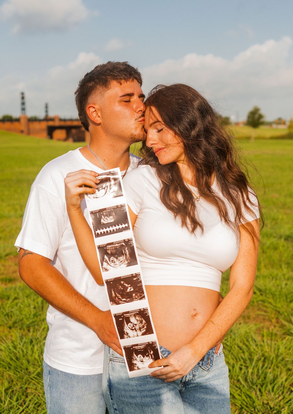 Couple in white outfits holding ultrasound photos outdoors, sharing a kiss during a pregnancy announcement photoshoot.