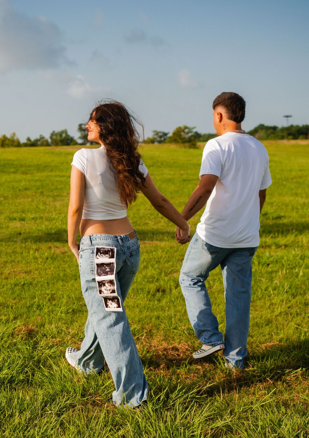 Couple in white tees and jeans holding hands in green field, woman displaying ultrasound photos on her jeans.