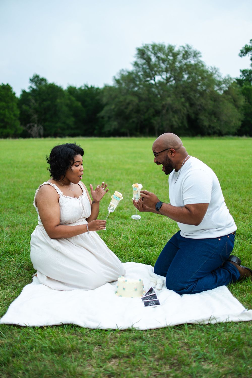 A couple plays cards together while sitting on a white blanket in a grassy field.