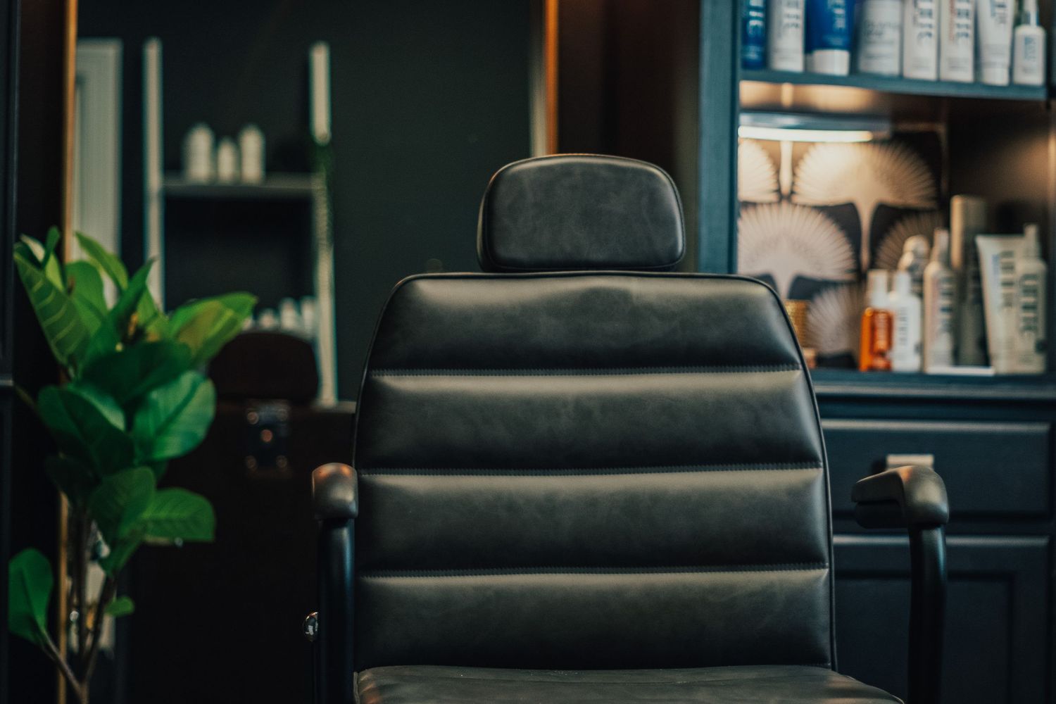 Black leather barber chair and potted plant in a modern salon setting with shelves of products in the background.