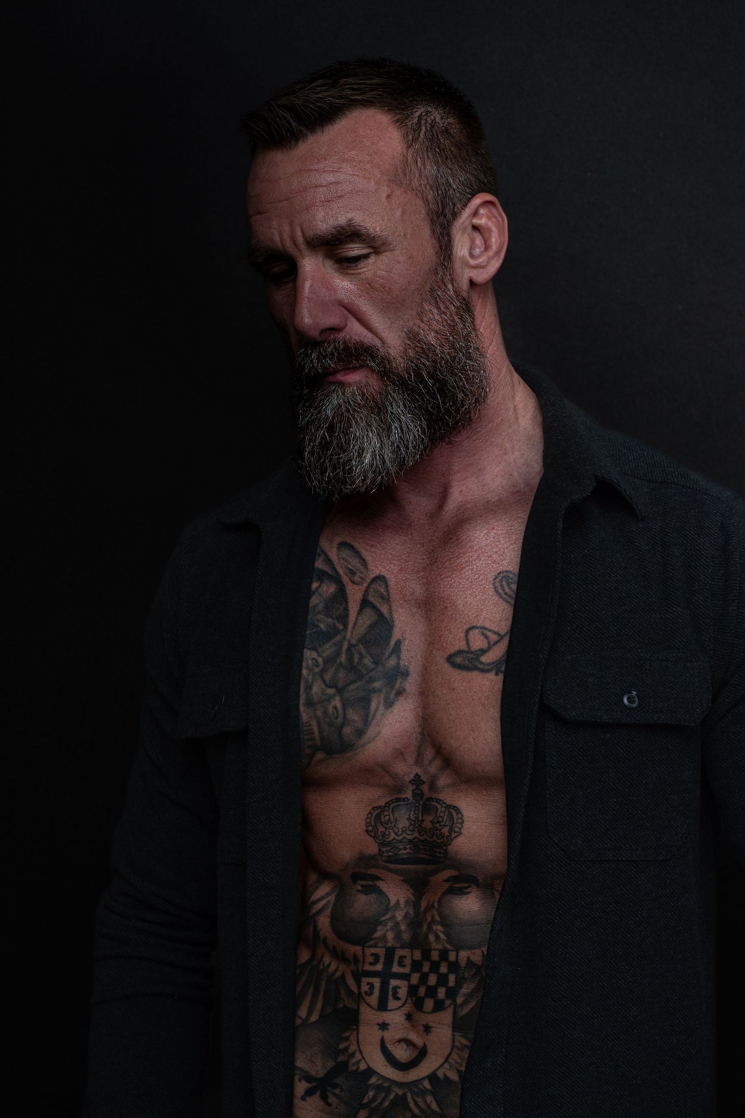 Dramatic dark portrait of muscular shirtless person with facial hair and elaborate chest tattoos wearing black shirt.