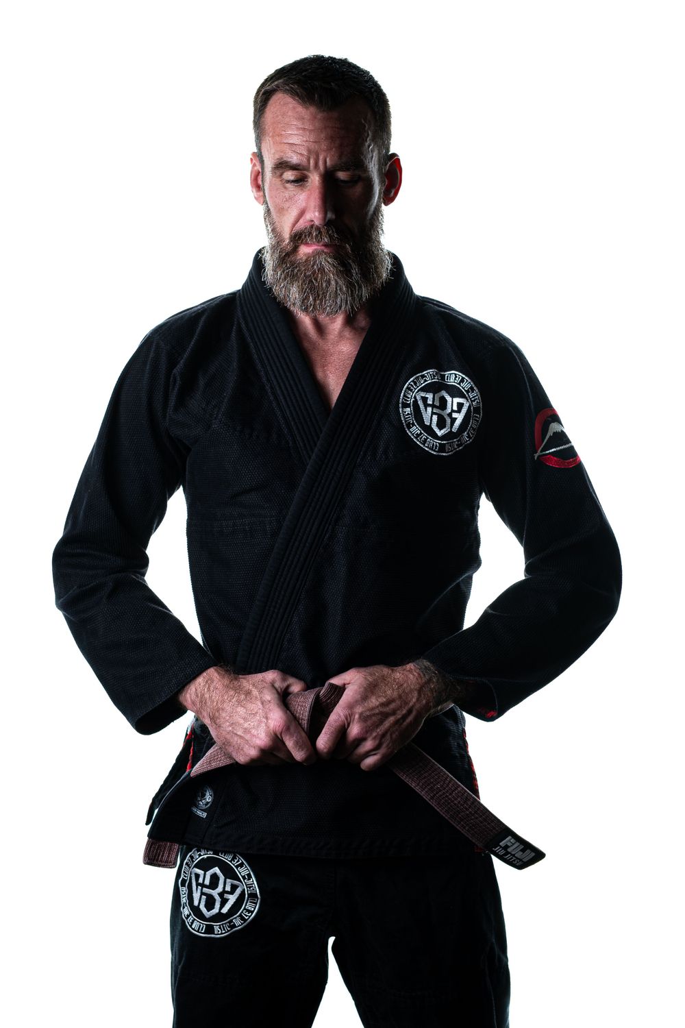 A martial artist wearing a black Brazilian Jiu-Jitsu gi with emblems and a brown belt stands in a strong pose.