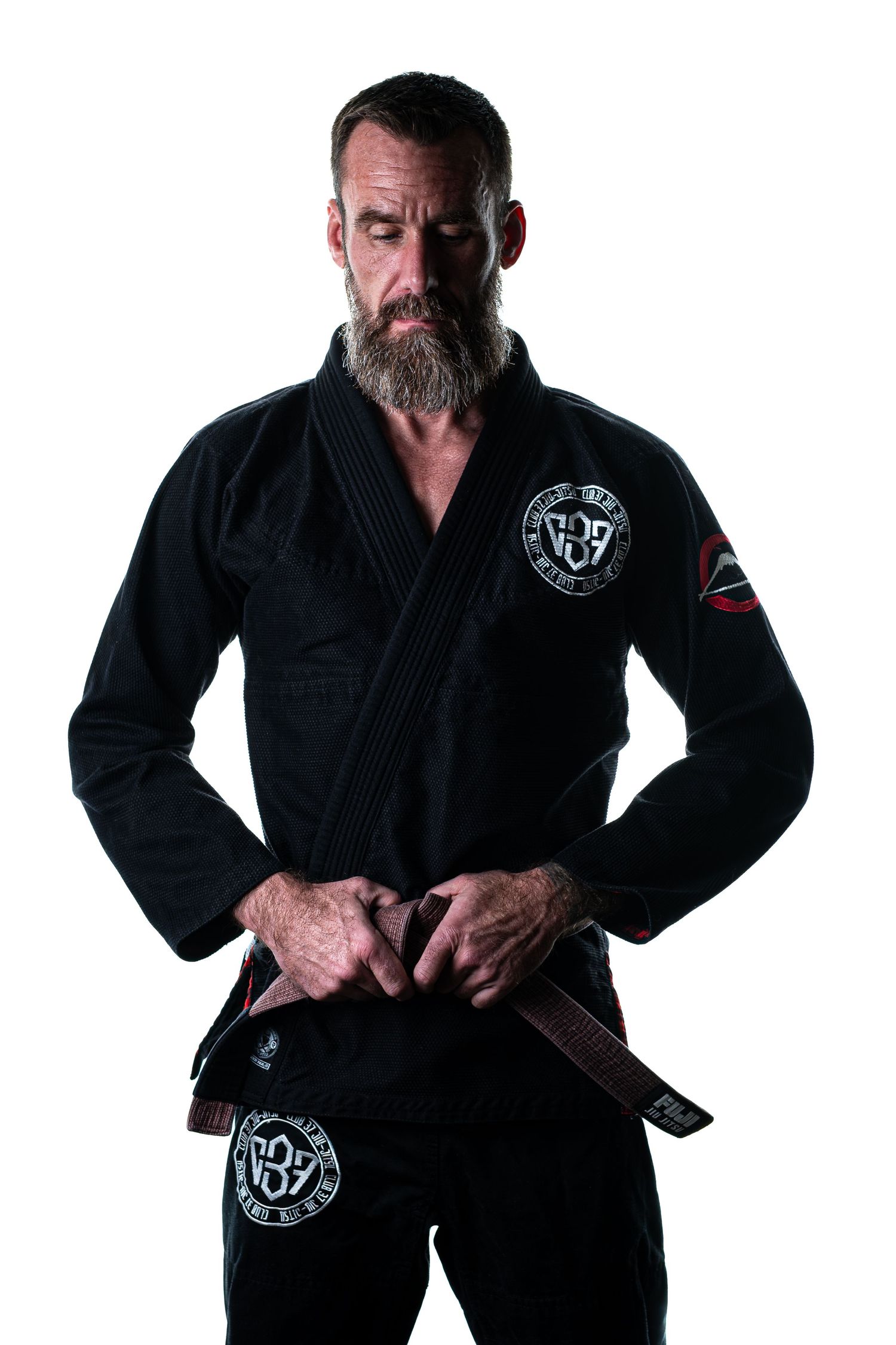 A martial artist wearing a black Brazilian Jiu-Jitsu gi with emblems and a brown belt stands in a strong pose.