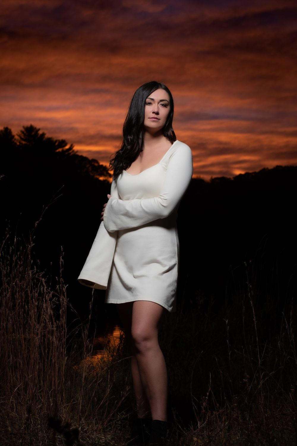 Artistic portrait of someone in a white sweater dress posing in a field at sunset with dramatic orange sky.