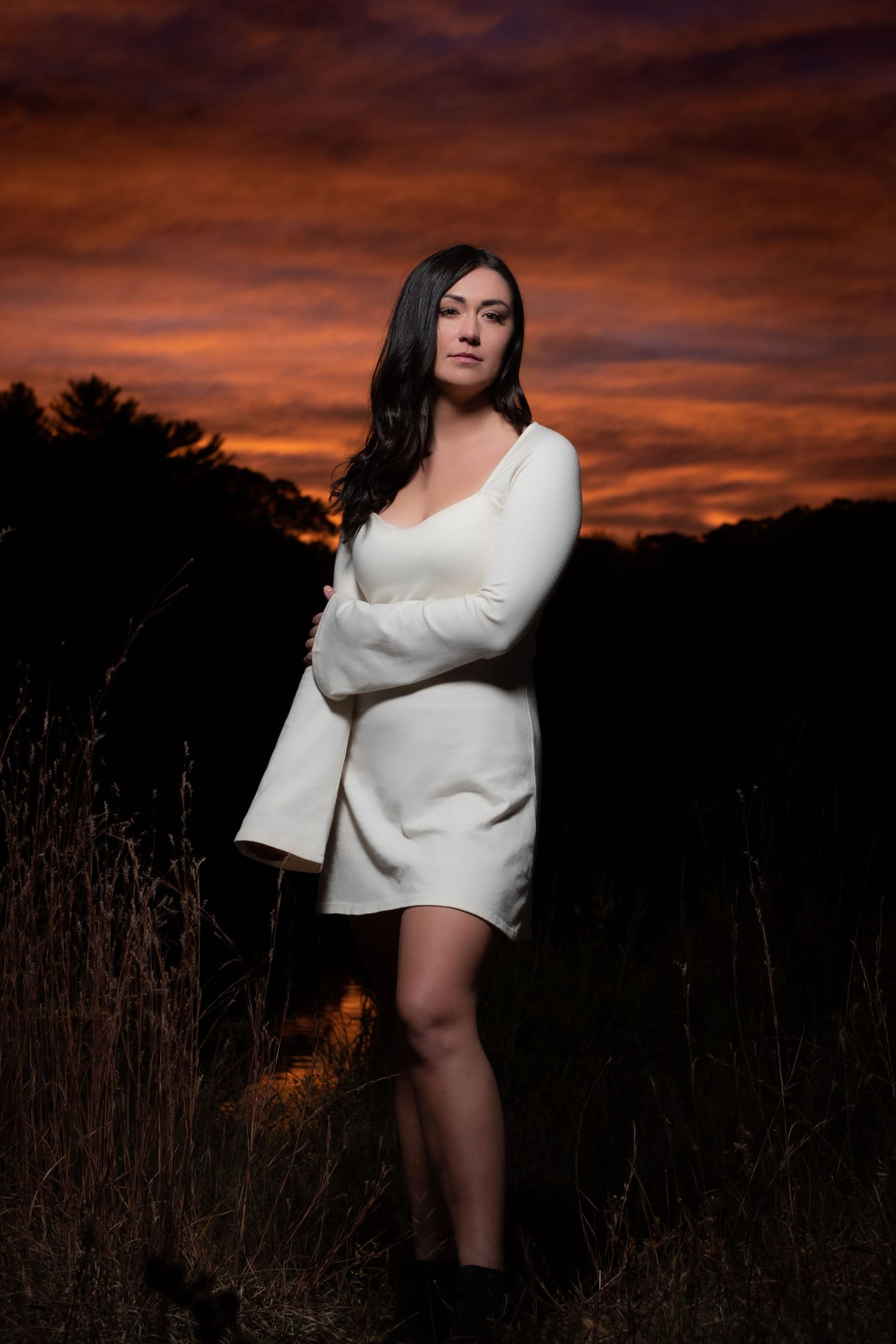 Artistic portrait of someone in a white sweater dress posing in a field at sunset with dramatic orange sky.