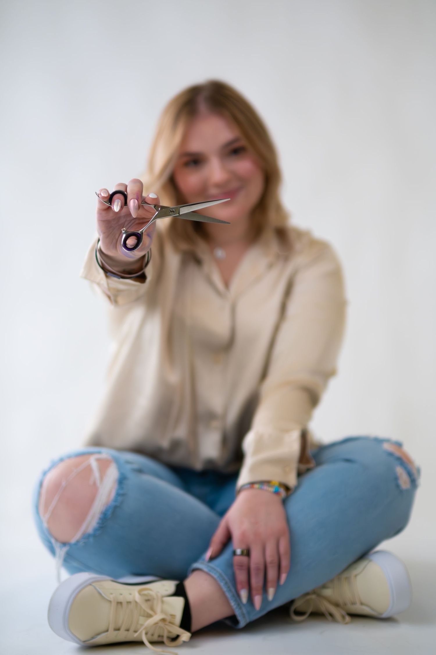 Someone sitting cross-legged on floor wearing ripped jeans and beige sweater holds up scissors against white background.