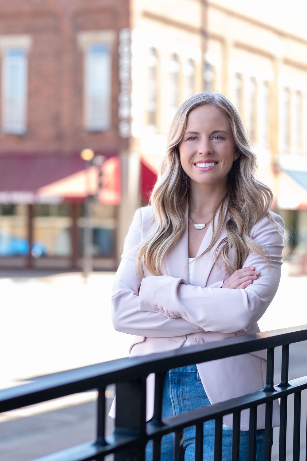 Professional portrait of a person in a light blazer and jeans standing by a railing in front of a downtown storefront.