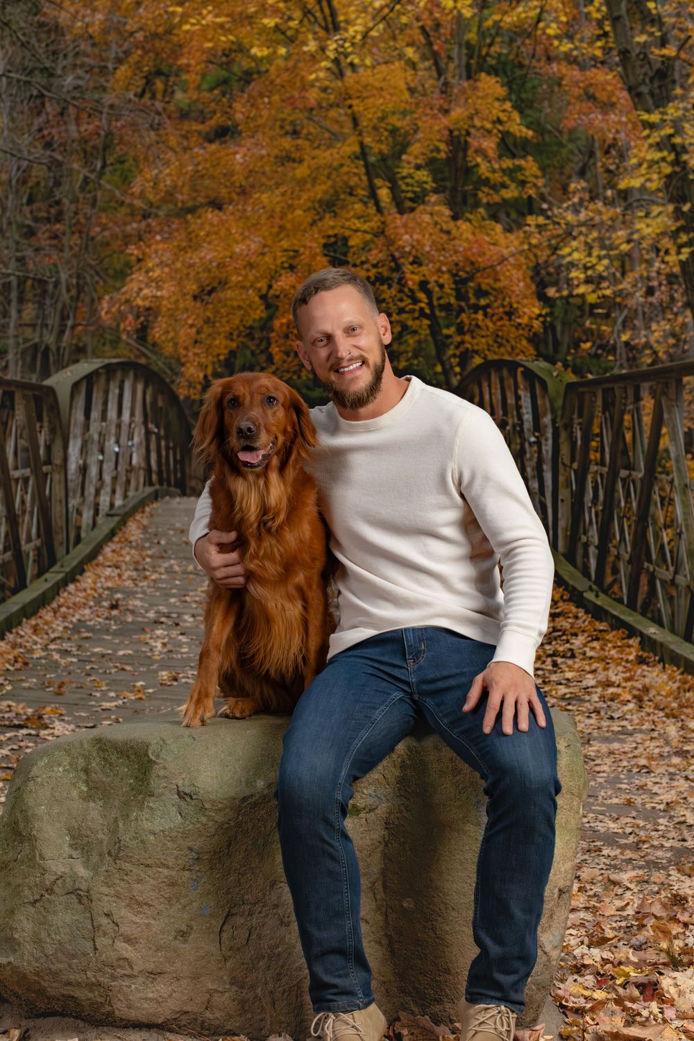 A beautiful autumn scene on a bridge with a Golden Retriever and person sitting on a rock amid colorful fall foliage.